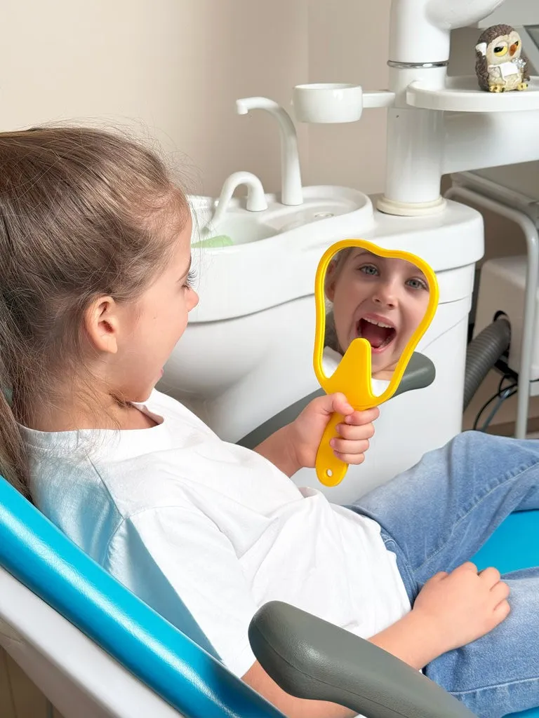 Lovely little girl looking in the mirror at her teeth after teeth surgery in a pediatric stomatology. A little girl sits in a dental chair and examines her cured teeth while looking in the mirror Lovely little girl looking in the mirror at her teeth after teeth surgery in a pediatric stomatology. A little girl sits in a dental chair and examines her cured teeth while looking in the mirror