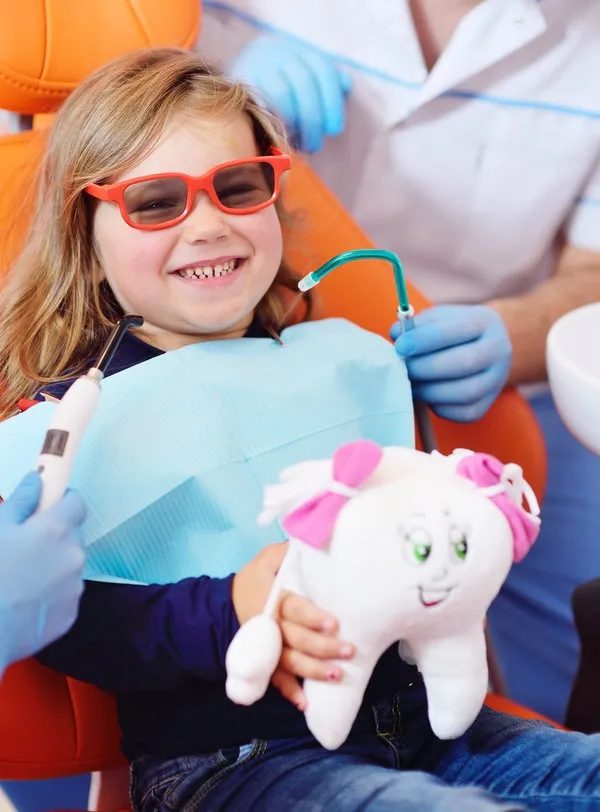 baby girl in an orange dental chair smiles while holding a soft toy in the shape of a tooth. The dentist holds in his hands an ultraviolet curing light for polymerization of the seal. A baby girl in an orange dental chair smiles while holding a soft toy in the shape of a tooth. The dentist holds in his hands an ultraviolet curing light for polymerization of the seal.