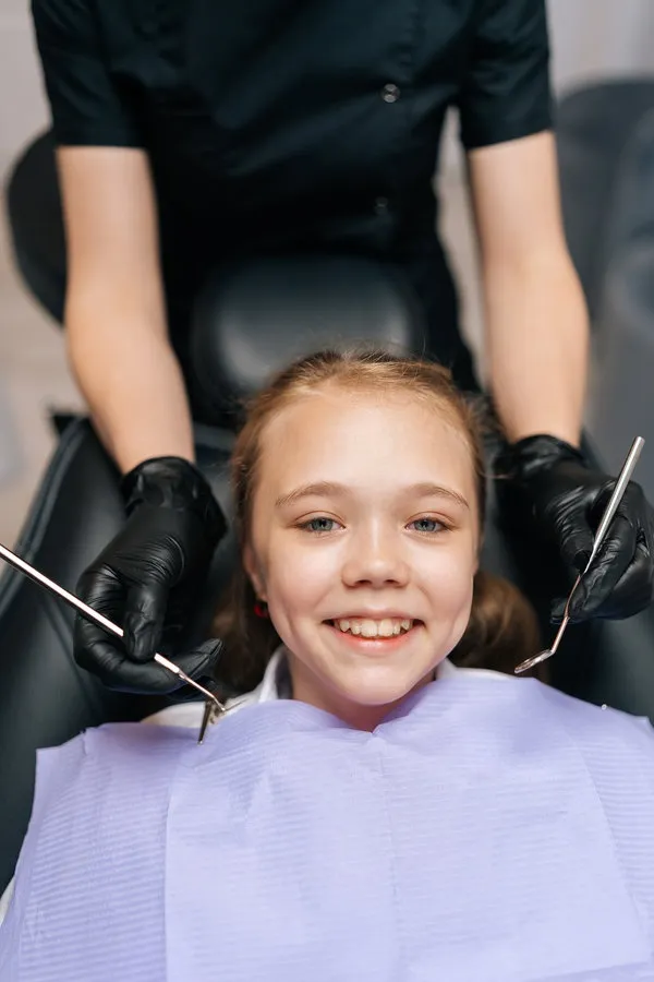 Vertical closeup portrait of happy cute little girl smiling looking at camera sitting in stomatology seat while pediatric dentist in gloves ready to do teeth examination. Concept of teeth treatment.