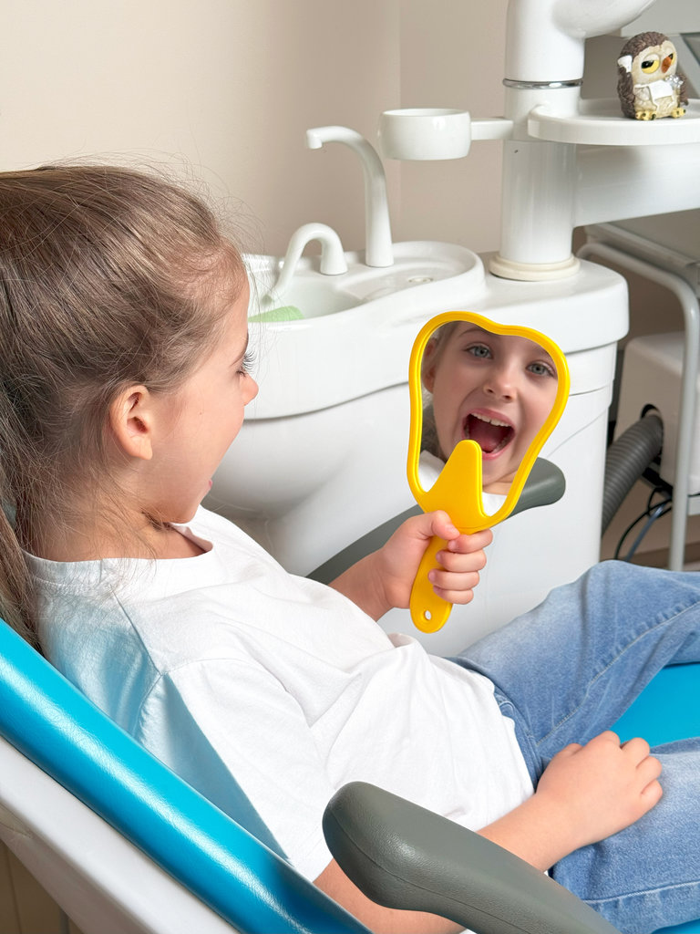 Lovely little girl looking in the mirror at her teeth after teeth surgery in a pediatric stomatology. A little girl sits in a dental chair and examines her cured teeth while looking in the mirror