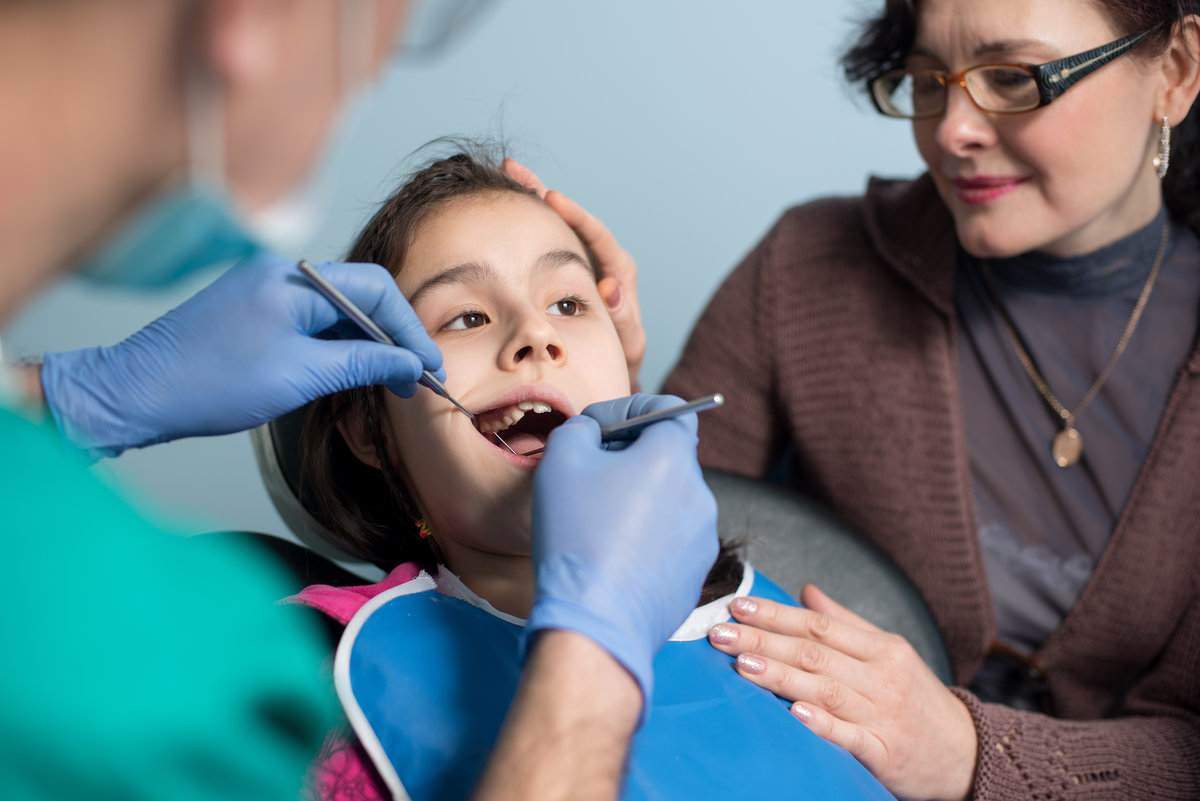 Young girl with mother on the first dental visit. Senior male dentist doing first check-up for patient at the dental office. Dentistry, medicine, stomatology and health care concept. Dental equipment