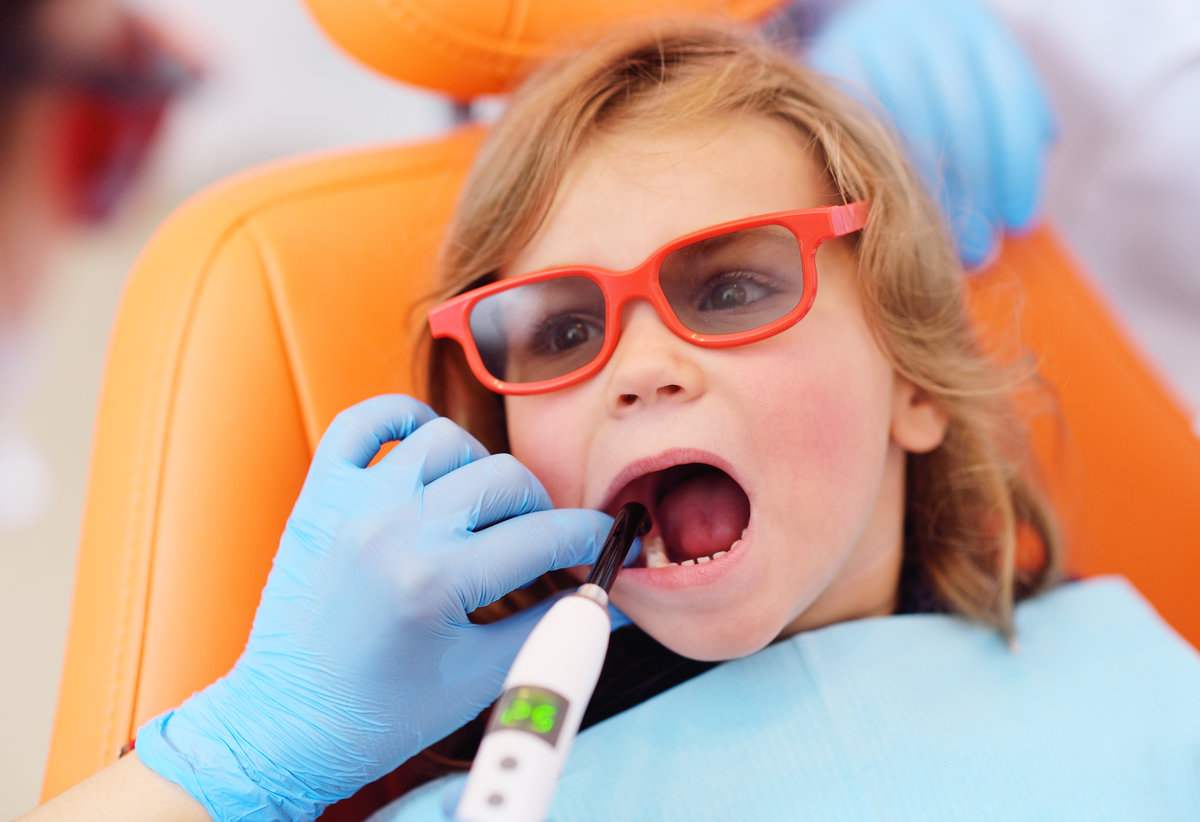 a pediatric dentist seals the teeth of a little girl in a pediatric dental clinic. A doctor using dental ultraviolet curing light to polymerize a seal