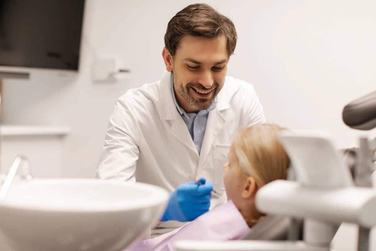 Male dentist talking to child girl sitting in dental chair in stomatology clinic, doctor preparing to start check-up