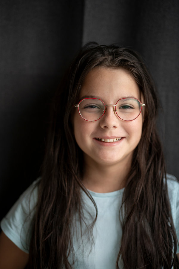 Smiling girl with long hair and glasses showing her teeth against a dark fabric background, soft light, copy space. Concept of dental health, confidence, and natural beauty. Smiling girl with long hair and glasses showing her teeth against a dark fabric background, soft light, copy space. Concept of dental health, confidence, and natural beauty.